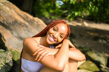 Obraz premium Outdoor portrait of natural Beautiful young African American woman with red braids hair style and perfect white teeth smile, posing in swimsuit at sunny summer day with rocks beach background