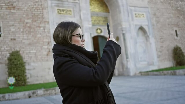 A brunette woman in glasses uses a smartphone at the topkapi palace in istanbul, evoking travel and history.