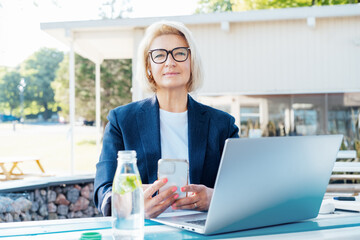 Smiling 50's stylish, confident mature business woman, middle aged lady using wireless ear buds, phone and laptop, working remotely in park. Caucasian woman make a video call, study, work online