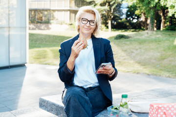 Middle-aged business woman eating sandwich burger with salad and using phone sitting on bench in the park while having lunch break. Remote work,business, freelance,blogging,social media concept