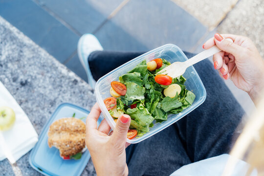 Top view close up lunch box with fresh salad and wooden folk in hand of woman sitting on the bench. Balanced diet lunchbox for weight loss. Healthy eating habits and well-being. Selective focus.
