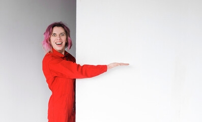 Your message is here. A stunned beautiful young woman in a red jumpsuit standing against a white wall points to the side