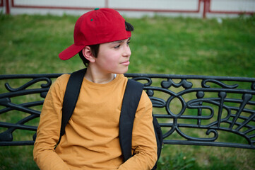Portrait of a happy teenage schoolboy in a red cap, sitting on a park bench, smiling and looking...