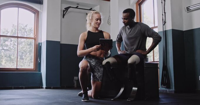 Young African Male double amputee with prosthetics talking to female personal trainer with a tablet in indoor fitness gym.