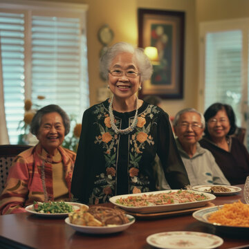 Joyous Elderly Woman Standing By The Dinner Table Between Her Seated Family Members Asian American.