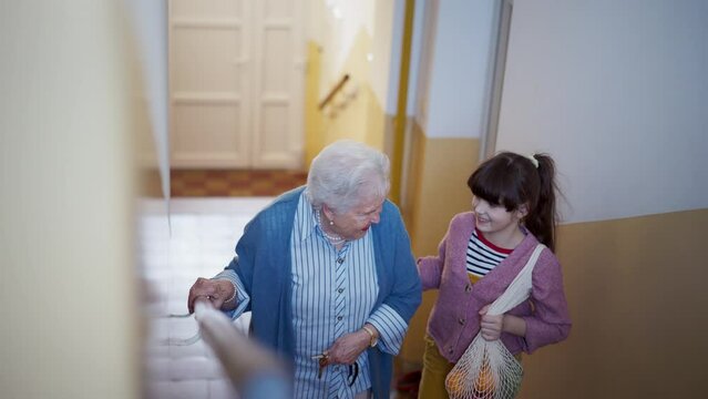 Granddaughter helping senior grandmother walk up the stairs, going grocery shopping. Girl holding hand of her elderly woman, providing stability while walking.