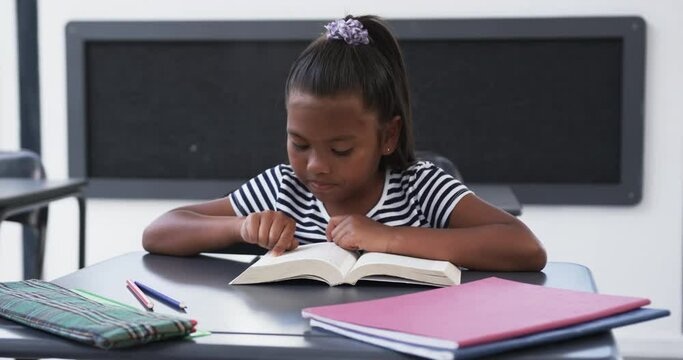 In school, in a classroom, a young African American girl is focused on reading a book