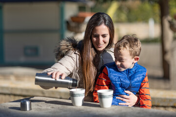 A smiling mother shares hot chocolate with her young son in a picnic area