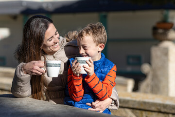 A smiling mother shares hot chocolate with her young son on a picnic area, while they look at each other knowingly.