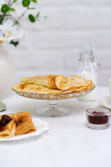 Thin pancakes with berries jam on glass stand with jam, bottle of milk, vase with flowers on white bricks background, top view.