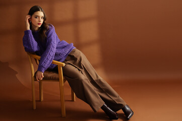 Fashionable confident beautiful brunette woman wearing trendy purple knitted sweater, pants, leather boots, posing on brown background. Studio fashion portrait. Copy, empty, blank space for text - 764213466
