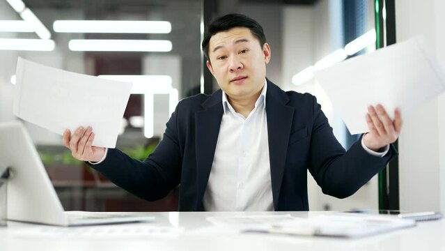 Confused asian businessman having difficulty with paper work sitting at workplace in business office. Puzzled mature man in a formal suit checks documents and cannot understand problem looks at camera