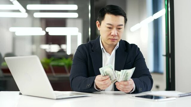 Serious asian businessman in a formal suit counting dollar bills while sitting at desk at workplace in business office. Focused mature employee holding cash in hands, calculates his earnings, salary