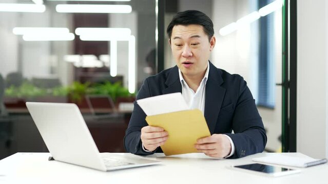 Happy joyful asian businessman reading letter with great news while sitting at workplace in business office. A man in a formal suit is celebrating success, pleased to receive a positive notification