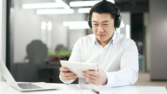Happy asian man in wireless headphones plays a computer game on a digital tablet while sitting at desk at workplace in business office. Excited smiling mature worker engrossed in interesting game