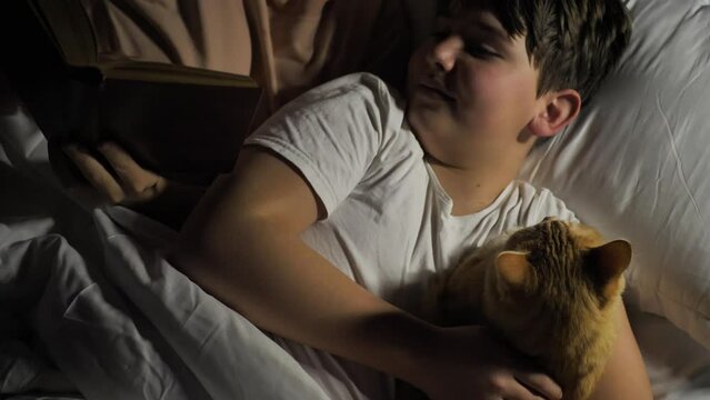 Mom reads a book to her son, lying together in bed with a beautiful ginger cat and getting ready for bed. Reading fairy tales to children before bed. Parental love and care. Friendship with pets
