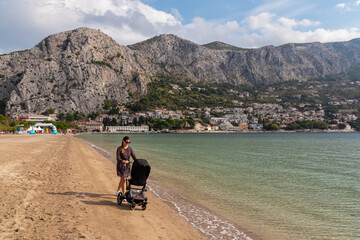 Loving mother with baby stroller walking on idyllic sand beach in coastal town Omis, Split-Dalmatia, South Croatia, Europe. Majestic coastline in Dinaric Alps, Adriatic Sea in Balkans. Family travel