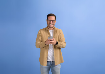 Portrait of cheerful male freelancer in glasses messaging client over smart phone on blue background