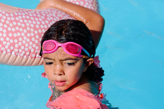 A girl is in the water with swimming goggles and pink swimsuit, holding on to a float and with a wet face. Vacation concept, time to relax and have fun.