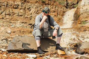 A man in shorts and a hat is leisurely sitting on a bedrock outcrop near a majestic waterfall in the valley, surrounded by stunning mountain landscape formation