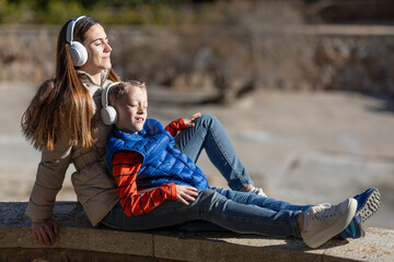 A mother and her son are sitting with a relaxed face, on a beach listening to music wih white headphones