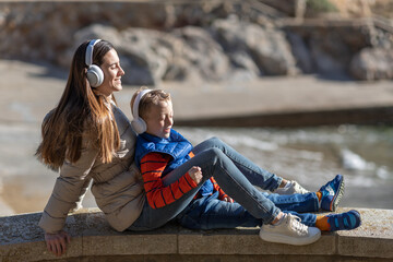  A mother and her son are sitting with a smiling face, on a beach listening to music with headphones