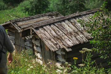 A man is standing in front of a rustic wooden shack surrounded by grass and natural landscape, giving a vintage feel to the scene