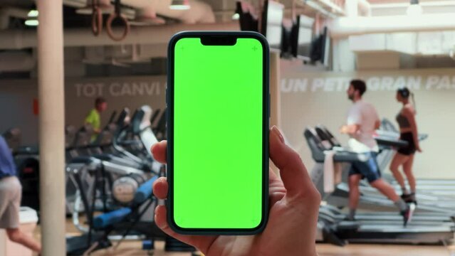 Woman using mobile phone while resting during fitness training. Hand holding smartphone with empty green screen chroma key at the gym.