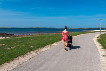 Loving mother walking with baby stroller along coastal road in Medulin, Istria peninsula, Croatia, Europe. Idyllic coastline of Kvarner Gulf in Adriatic Mediterranean Sea. Travel destination in summer