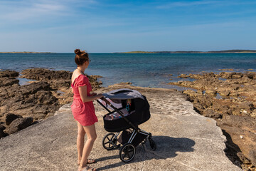 Loving mother walking with baby stroller along coastal road in Medulin, Istria peninsula, Croatia, Europe. Idyllic coastline of Kvarner Gulf in Adriatic Mediterranean Sea. Travel destination in summer