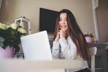 Young Beautiful Asian business woman working online with laptop computer placed at the table at home office. Smiling lady freelancer have remote work sitting in living room with white roses flowers