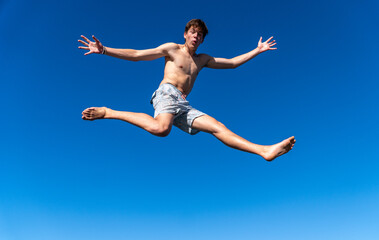 A joyful young man leaps in the air with arms wide open and a surprised expression, against a backdrop of beautiful blue sky on a sunny summer day.