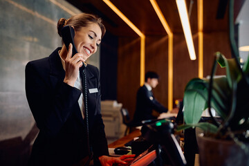 Happy female receptionist answering phone call while working at hotel front desk.
