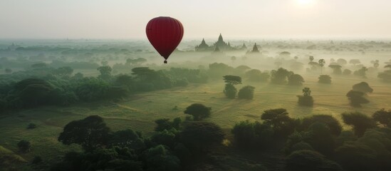 A red hot air balloon is flying over a lush green field. The sky is hazy and the sun is setting - obrazy, fototapety, plakaty