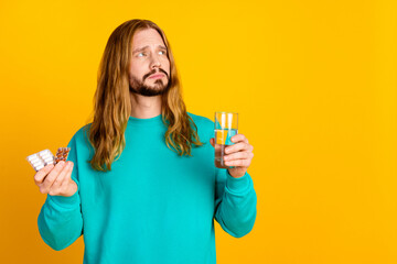 Portrait of unsatisfied minded guy hold pills glass water look empty space isolated on yellow color...