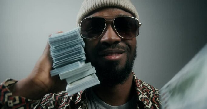 A bearded man in a hat and sunglasses, with a wad of money in his hand, smiling at the camera standing on a gray background in the studio. Dollar bills are falling on top of him