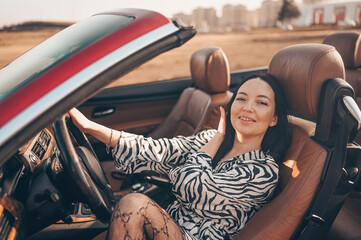 Happy smiling brunette woman driver sitting in new red cabriolet car on beach coast, smiling...