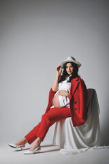 Stunning brunette pregnant woman in white hat, shirt and red suit posing on a cyclorama. Copy space for text