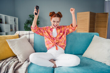 Full size portrait of delighted girl sit sofa raise fist hold tv remote control shout yeah living...