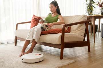 Young asian woman relaxing with pug dog in living room while Robotic vacuum cleaner working.