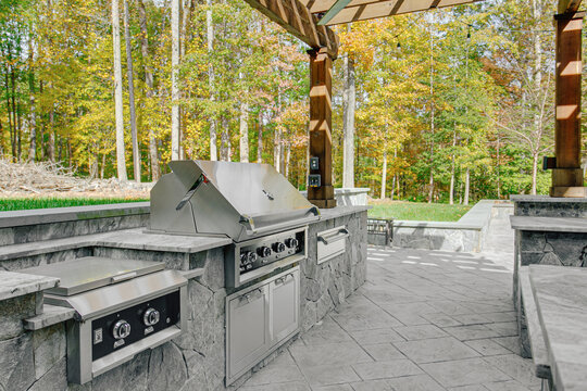 Inviting Partially Covered Outdoor Patio with Large Stone Kitchen Island, Grill, and Seating Area Framed by Wooden Pergola Amidst Vibrant Foliage