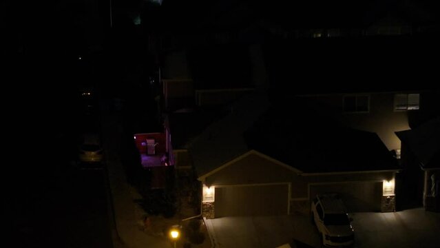 Aerial view at night of the side yard and patio of a condo with colorful landscape lighting and garage and condominium roofs partially covered with snow.