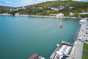 A picturesque aerial view of the mountain lake with a tour boat on the water, the embankment with boats and catamarans moored to the shore against the background of the resort architecture.
