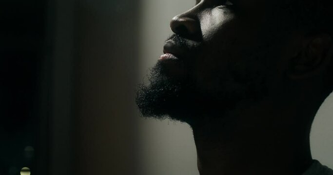 A bearded man in casual clothes leaned his head against the wall with a depressed look, close-up of his face. Dim indoor lighting