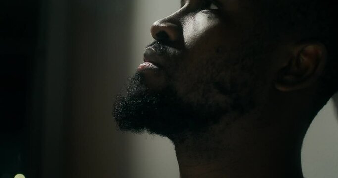 A bearded man in casual clothes leaned his head against the wall with a depressed look, close-up of his face. Dim indoor lighting
