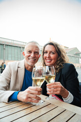 Vertical. Mature couple toasting white wine glasses smiling and looking at camera. Middle aged married people drinking alcohol on a bar terrace. Mid adult friends clinking with alcohol beverages