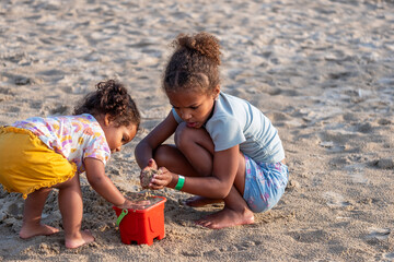 Two young African American children, absorbed in filling a bucket with sand, share a moment of cooperative play on the beach, sibling love and National Siblings day.