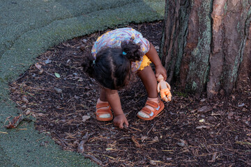 A little mixed race child bends down to touch the ground near a tree, immersed in play and natural...