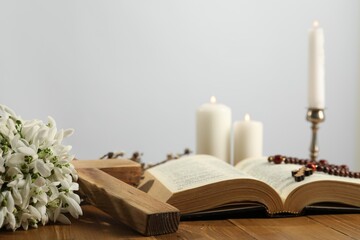 Church candles, cross, rosary beads, Bible and flowers on wooden table against light background