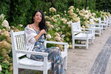 A pensive woman in a floral dress lounges on a white garden bench, her shawl draped casually,...
